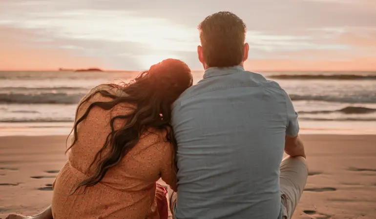 a man and woman sitting on the beach watching the sunset