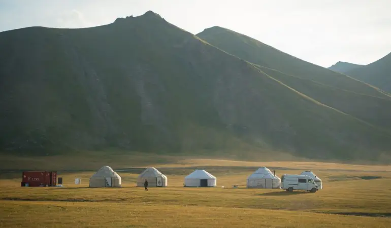 Row of yurts and camper van at dawn with scenic mountain backdrop, serene travel setting.