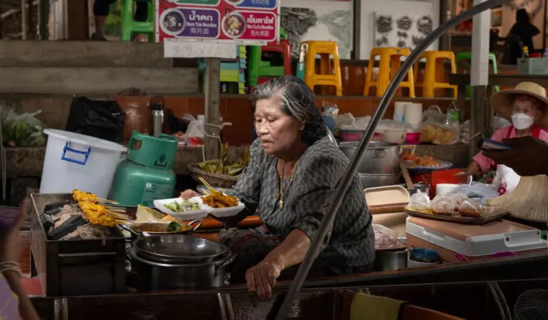 Elderly woman cooking traditional Thai dishes at a bustling Bangkok street market.