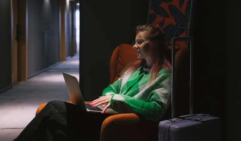 A woman in a hotel hallway using a laptop, sitting on an armchair next to luggage, creating a cozy working environment.